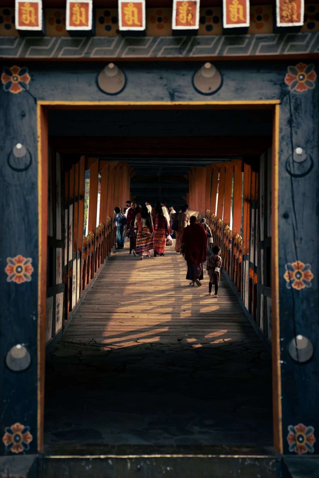 A monk in maroon robes and a child walking with others along a sunlit covered wooden bridge