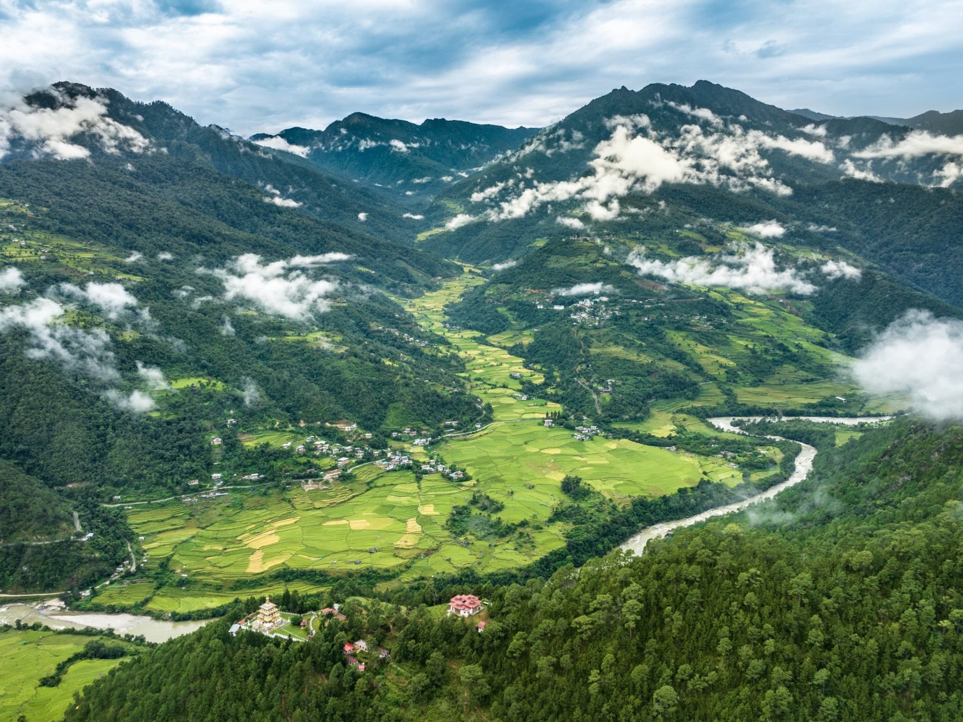 An aerial view of a remote Bhutanese valley with terraced rice fields, a winding river, and a small monastery rising from the forest floor