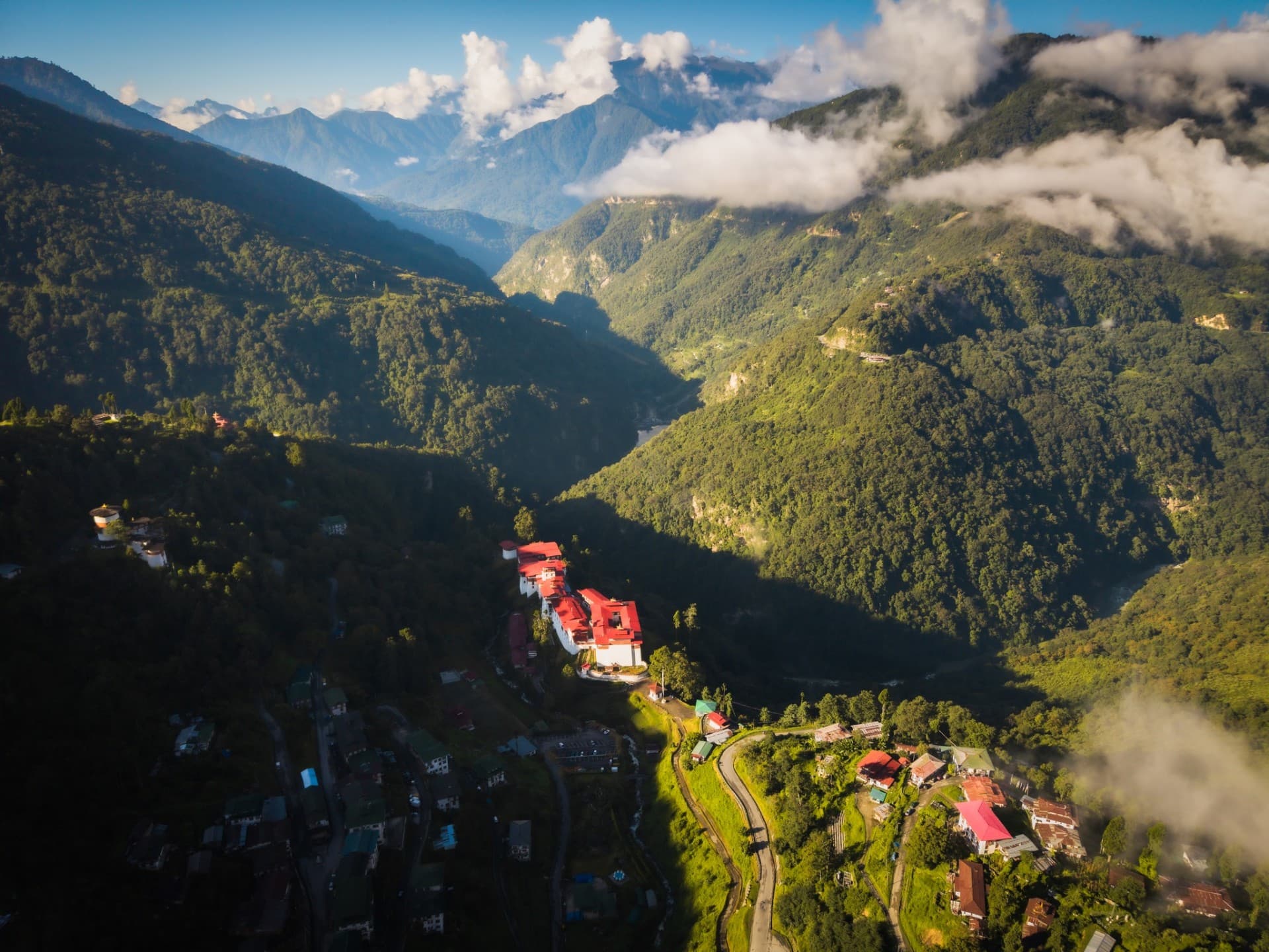 Aerial view of a Bhutanese dzong on a forested ridge with the Himalayas rising through cloud