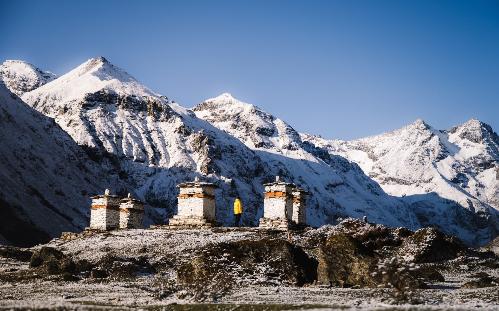 A solitary figure in a yellow jacket standing among three chortens beneath the snow-clad Bhutanese Himalaya