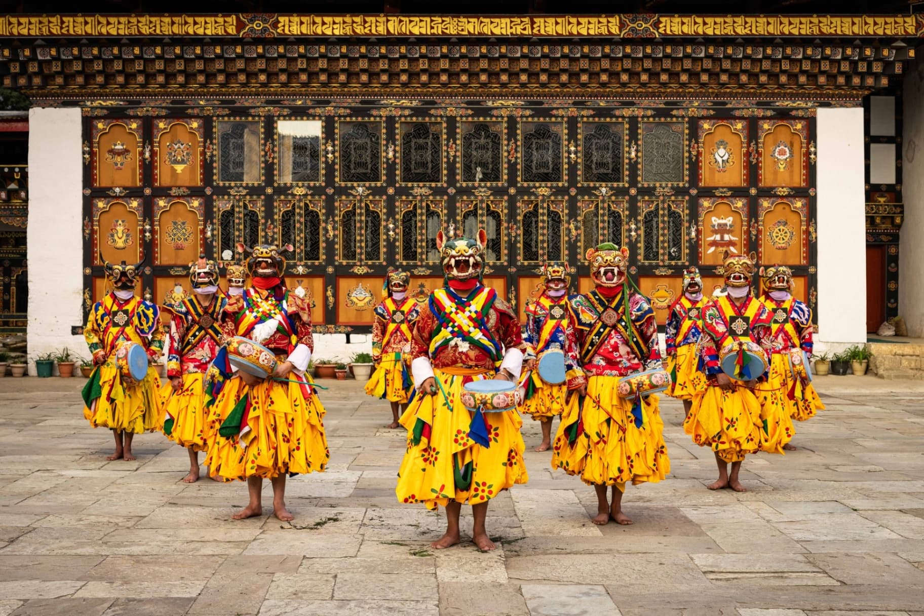 Masked Cham dancers in saffron robes performing before the painted facade of a Bhutanese dzong