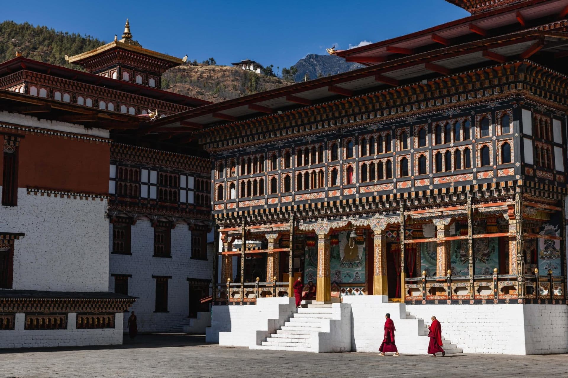 Two monks in crimson robes crossing the painted courtyard of a Bhutanese dzong beneath a bright Himalayan sky