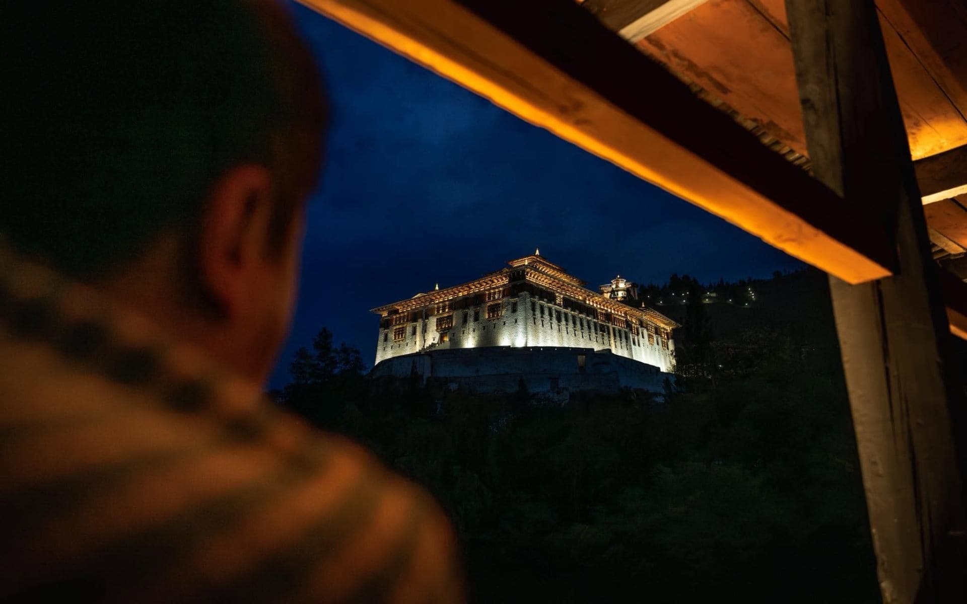 A monk gazes from a wooden bridge toward a Bhutanese dzong illuminated against the deep blue dusk sky