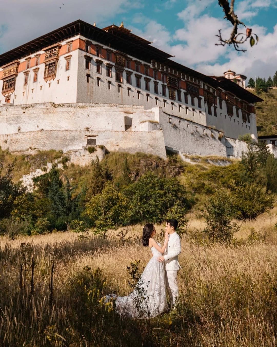 A couple in white in a meadow of golden grasses beneath the white walls of Rinpung Dzong, Paro