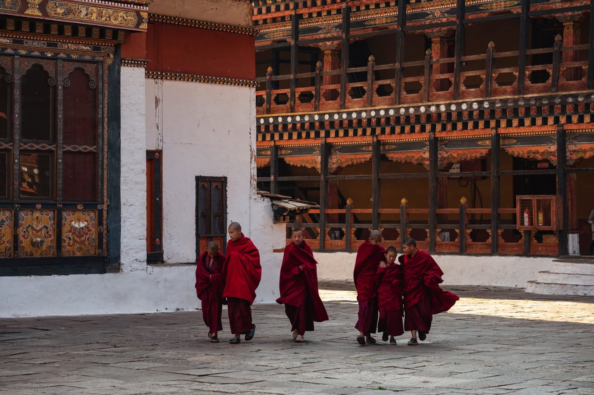 Young monks in crimson robes crossing the painted stone courtyard of Punakha Dzong in late afternoon light